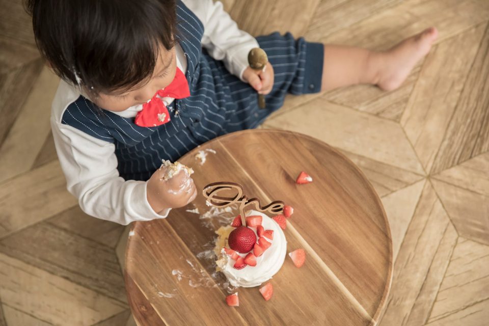 スマッシュケーキ: 蝶ネクタイを付けた一歳児がOneと書かれた飾りの付いた苺のケーキをつかみ食べしている